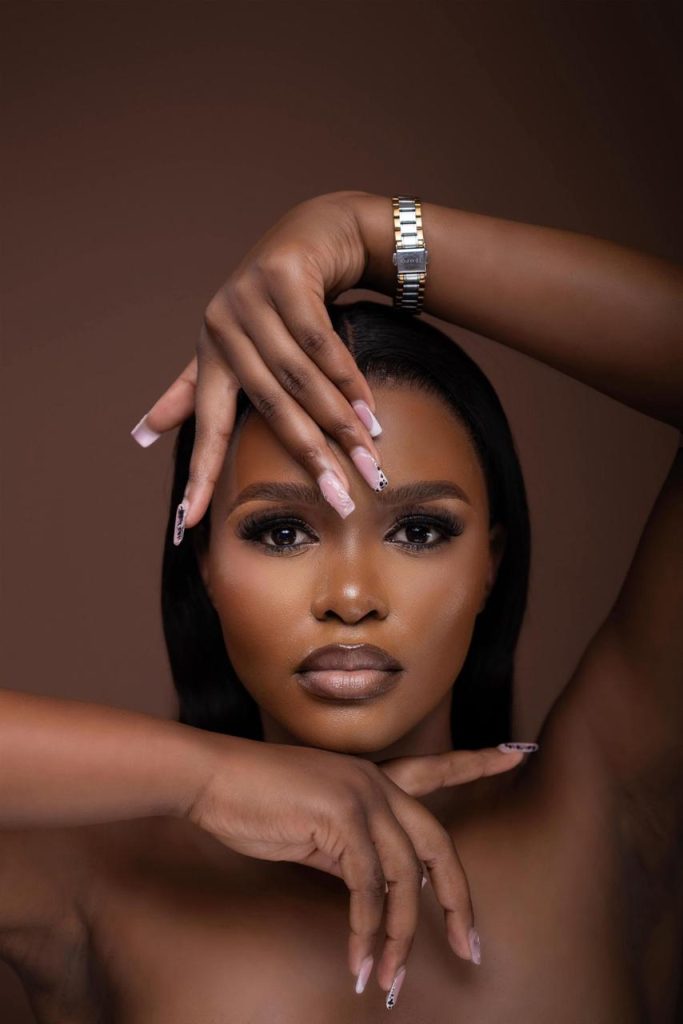 Close-up portrait of a confident Emihle with long dark hair, showcasing a striking facial expression and hands positioned elegantly around her face, set against a neutral background.